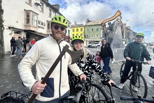 Cyclists with helmets and an axe pose near a large harp statue in a town square.