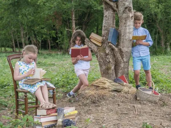 Three children reading books by a tree in a grassy area.